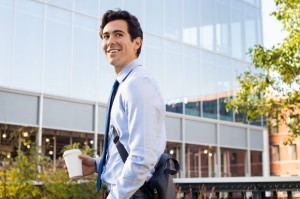 50076654 - happy young businessman walking and holding laptop bag and coffee paper cup. satisfied businessman looking away with modern buildings in background. happy smiling man going to work with a take away coffee in a paper cup.