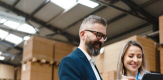 Starting a Logistics Company? Remember These Tips A professionally dressed man and woman stand in a storage warehouse and reference a tablet, both of them smiling.