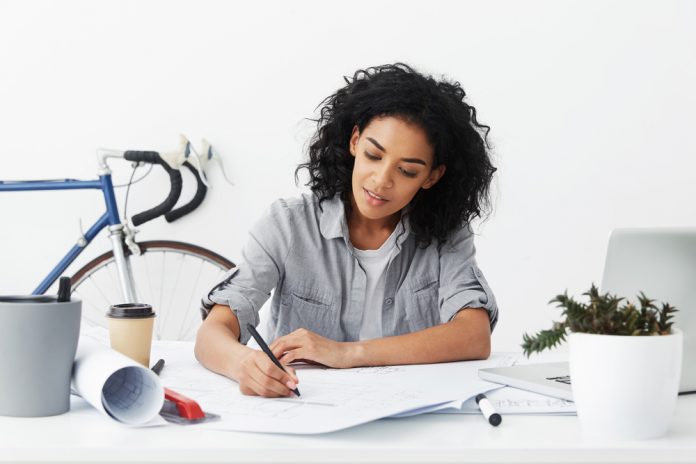 Smiling young self-employed woman architect making drawing at her home office using pen, excited about working process