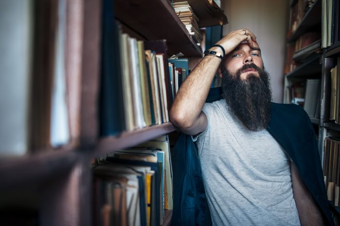 Thoughtful hipster man among bookshelves. Depression concept