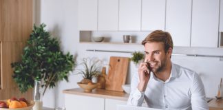 Smiling young businessman talking on smartphone during breakfast at kitchen table with laptop and coffee cup