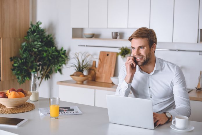 Smiling young businessman talking on smartphone during breakfast at kitchen table with laptop and coffee cup