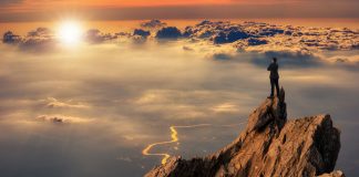 Businessman wearing comfortable casual suit jacket standing on top of peak mountain and looking forward