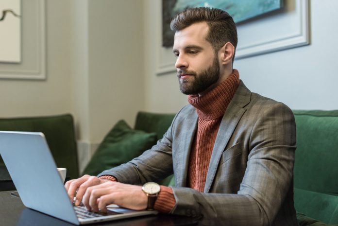 Handsome bearded businessman in formal wear sitting at table and typing on laptop in restaurant