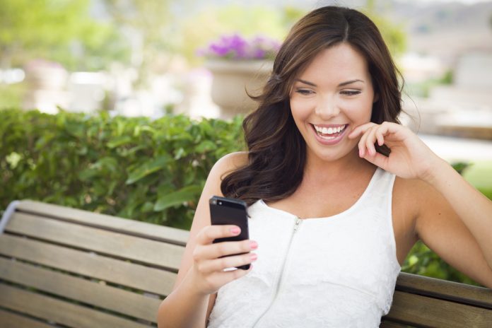 Smiling Young Adult Female Texting on Cell Phone Outdoors on a Bench