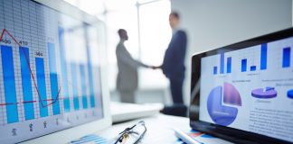 Eyeglasses, pen, two touchpads and financial documents at workplace with businessmen handshaking on background