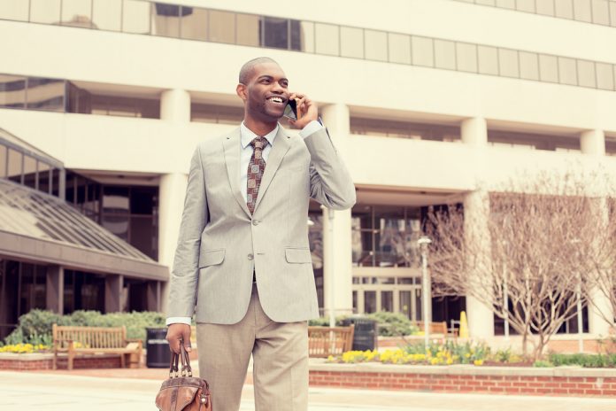 Young busy happy, smiling business man talking on mobile phone holding briefcase walking down the street outside corporate office isolated on city background