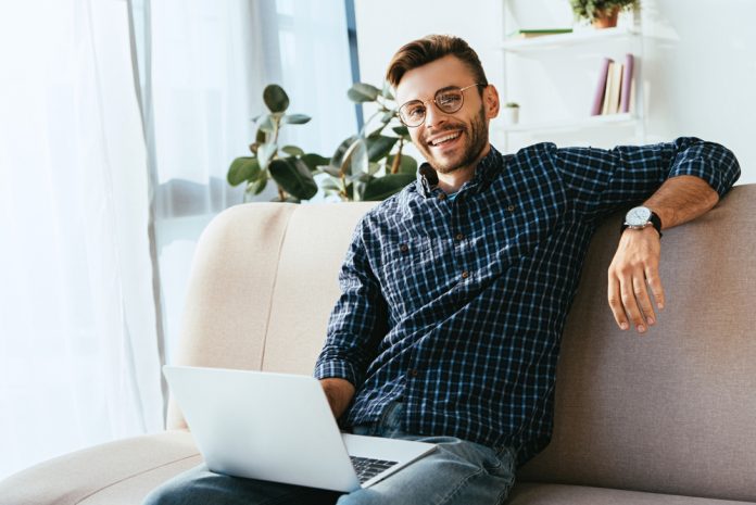 Smiling man in eyeglasses with laptop sitting on sofa at home