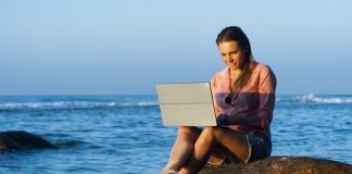 Woman typing on laptop while sitting on a rock above the ocean