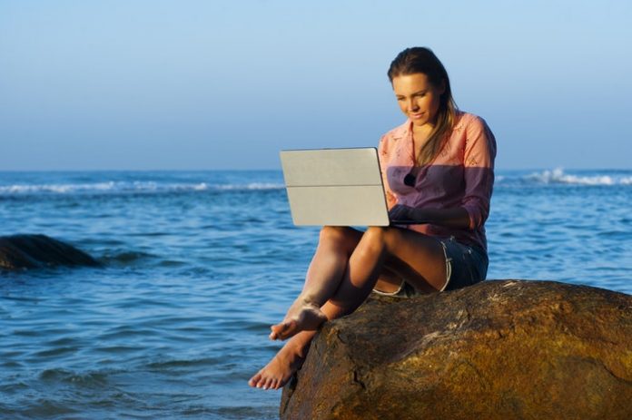 Woman typing on laptop while sitting on a rock above the ocean