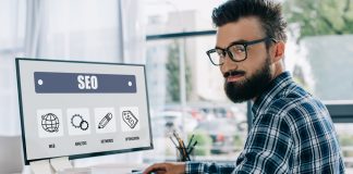 Young successful SEO manager sitting at workplace with computer and looking at camera