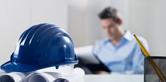 Hardhat and blueprint on desk, with businessman in background
