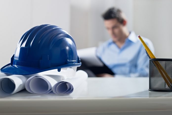 Hardhat and blueprint on desk, with businessman in background