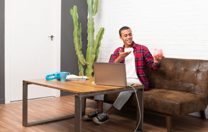 Man with laptop in living room holding a big piggybank