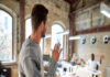 A young person walking out of a small work office and waving at a coworker who is seated at a white desk.
