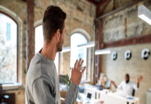 A young person walking out of a small work office and waving at a coworker who is seated at a white desk.
