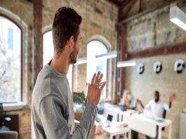 A young person walking out of a small work office and waving at a coworker who is seated at a white desk.