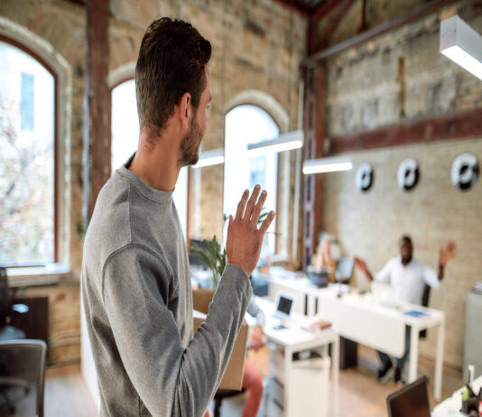 A young person walking out of a small work office and waving at a coworker who is seated at a white desk.