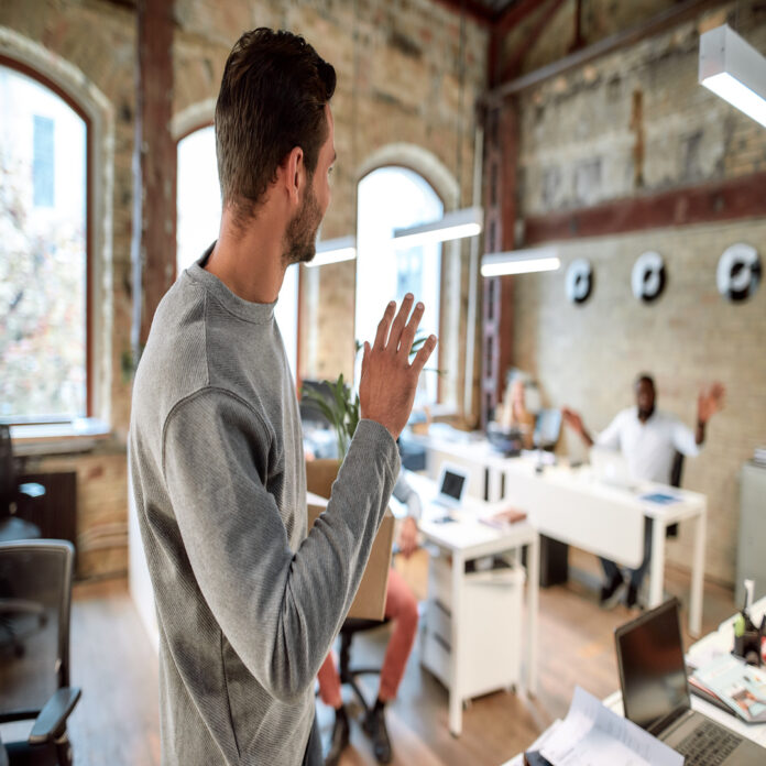 A young person walking out of a small work office and waving at a coworker who is seated at a white desk.