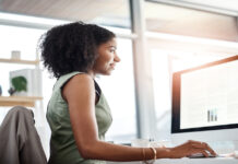 A woman in a green dress uses a mouse while reviewing data tables on a monitor in a sunlit office with large windows.