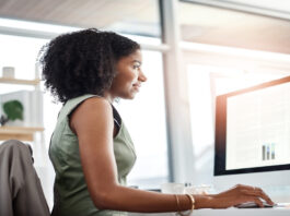 A woman in a green dress uses a mouse while reviewing data tables on a monitor in a sunlit office with large windows.