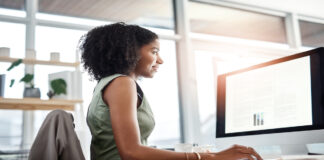 A woman in a green dress uses a mouse while reviewing data tables on a monitor in a sunlit office with large windows.