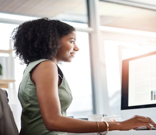 A woman in a green dress uses a mouse while reviewing data tables on a monitor in a sunlit office with large windows.