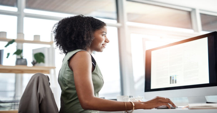 A woman in a green dress uses a mouse while reviewing data tables on a monitor in a sunlit office with large windows.