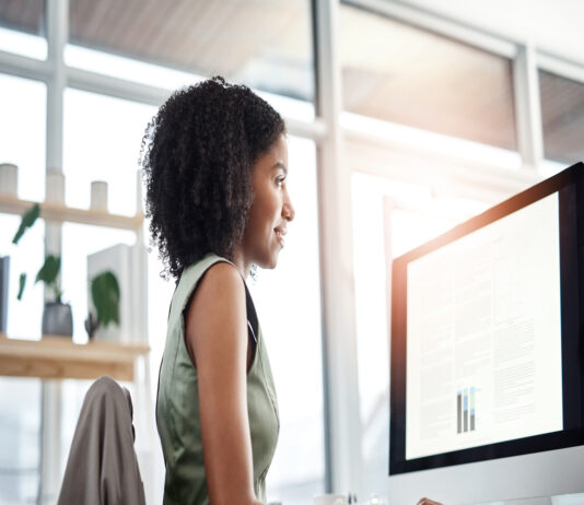A woman in a green dress uses a mouse while reviewing data tables on a monitor in a sunlit office with large windows.