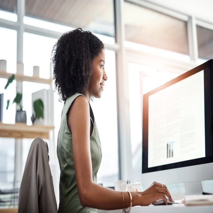 A woman in a green dress uses a mouse while reviewing data tables on a monitor in a sunlit office with large windows.