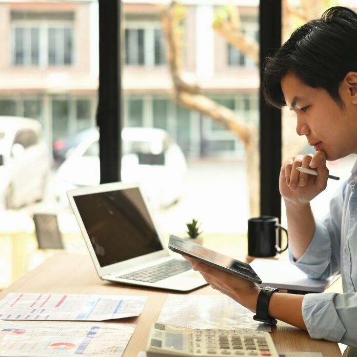 A professionally dressed man sits at a wooden desk holding a tablet in one hand and resting his chin on his other hand.
