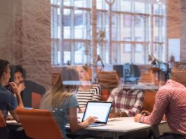 A startup team sits in a conference room with orange chairs and glass walls. They're all at the table talking.