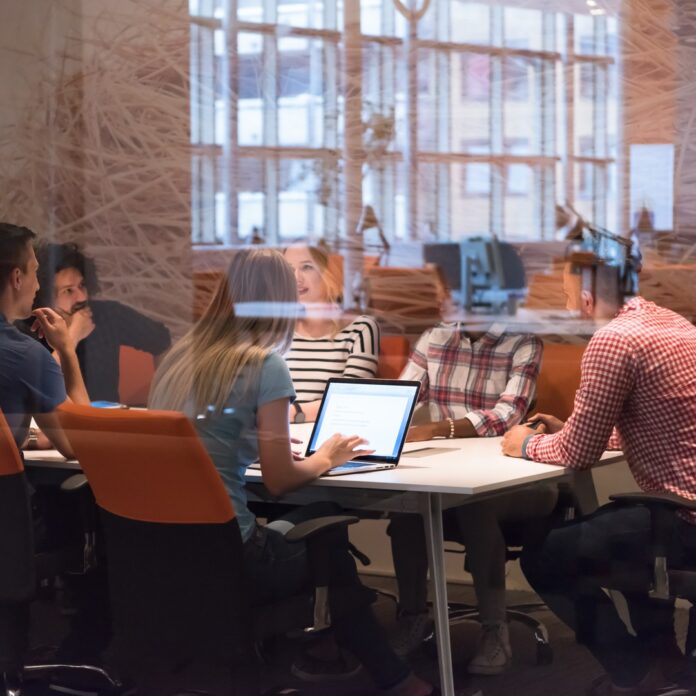 A startup team sits in a conference room with orange chairs and glass walls. They're all at the table talking.