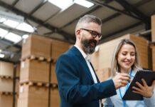 A professionally dressed man and woman stand in a storage warehouse and reference a tablet, both of them smiling.