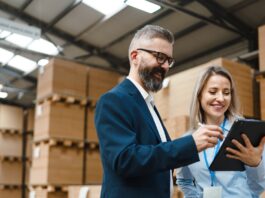 A professionally dressed man and woman stand in a storage warehouse and reference a tablet, both of them smiling.