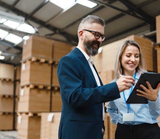 A professionally dressed man and woman stand in a storage warehouse and reference a tablet, both of them smiling.