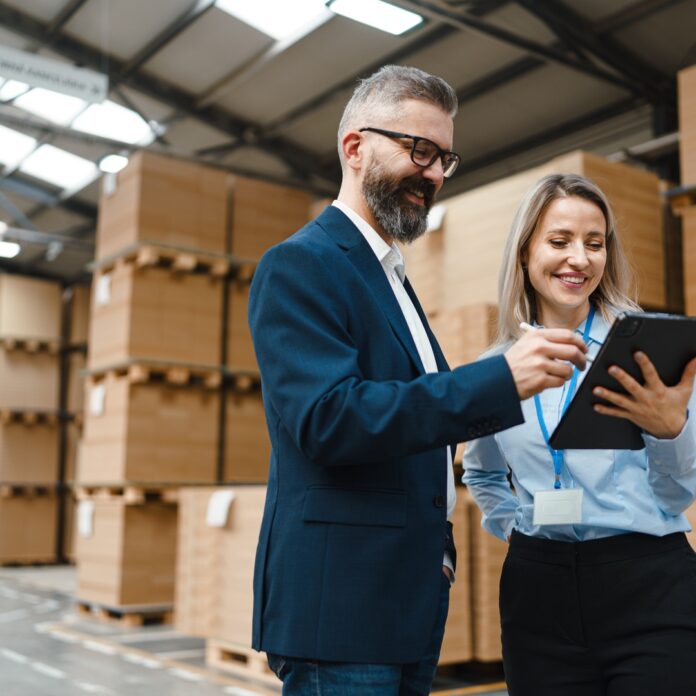 A professionally dressed man and woman stand in a storage warehouse and reference a tablet, both of them smiling. A professionally dressed man and woman stand in a storage warehouse and reference a tablet, both of them smiling.