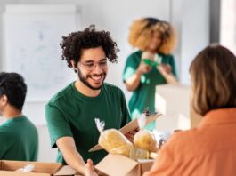 A man in a green shirt and glasses smiles as a woman in an orange shirt hands him a box of donated goods.