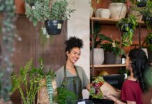 Why Interior Condition Impacts Customer Retention Florist smiles behind a shop counter, handing a wrapped bouquet to a customer among potted plants.
