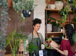 Florist smiles behind a shop counter, handing a wrapped bouquet to a customer among potted plants.