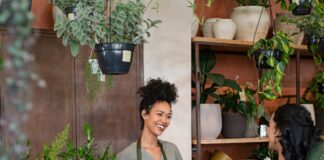 Florist smiles behind a shop counter, handing a wrapped bouquet to a customer among potted plants.