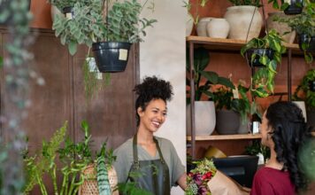 Why Interior Condition Impacts Customer Retention Florist smiles behind a shop counter, handing a wrapped bouquet to a customer among potted plants.