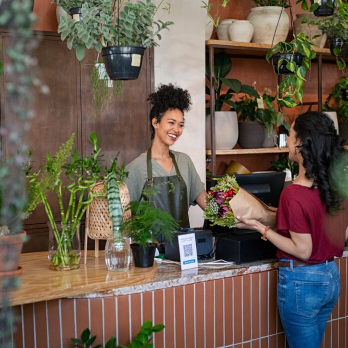 Why Interior Condition Impacts Customer Retention Florist smiles behind a shop counter, handing a wrapped bouquet to a customer among potted plants.