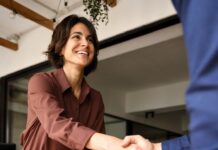 A businesswoman shake hands with a client across a desk during an office meeting, with papers and a laptop nearby.