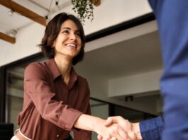 A businesswoman shake hands with a client across a desk during an office meeting, with papers and a laptop nearby.