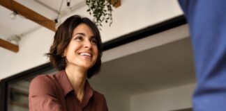 A businesswoman shake hands with a client across a desk during an office meeting, with papers and a laptop nearby.