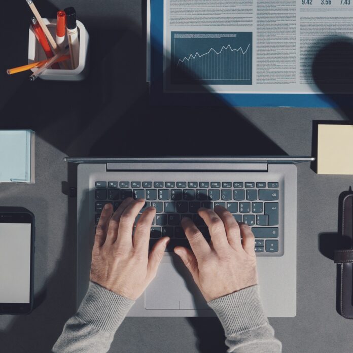 An aerial view of someone sitting at their desk with their hands on the laptop in the center and office supplies around them. An aerial view of someone sitting at their desk with their hands on the laptop in the center and office supplies around them.