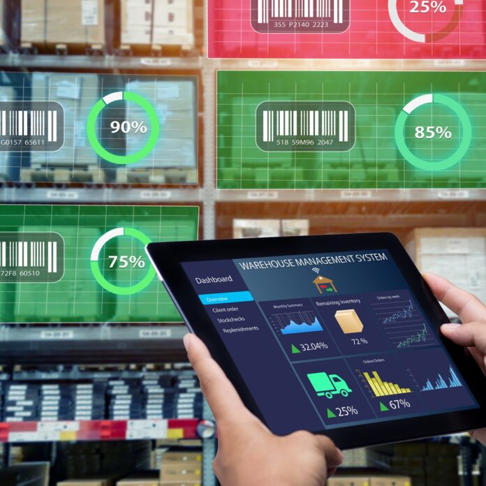 A worker holds a tablet showing warehouse data while shelves of goods display digital inventory tracking metrics. A worker holds a tablet showing warehouse data while shelves of goods display digital inventory tracking metrics.