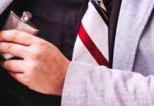 Screening Tools To Prevent Employee Substance Use at Work A close-up of a man’s chest wearing a black shirt and a striped tie, hiding a small flask under his grey suit jacket.