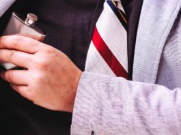 A close-up of a man’s chest wearing a black shirt and a striped tie, hiding a small flask under his grey suit jacket.